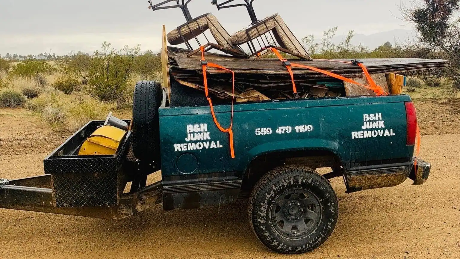 The B&H Junk Removal pickup loaded with old boards and two bar stools on open desert ground