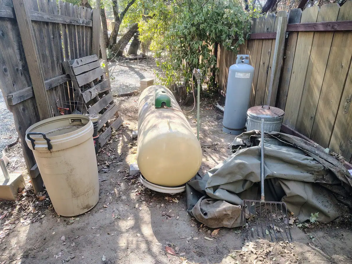 A side yard before the work: trash cans; broken pallets; a folded tarp and a pitchfork crowded along the fence