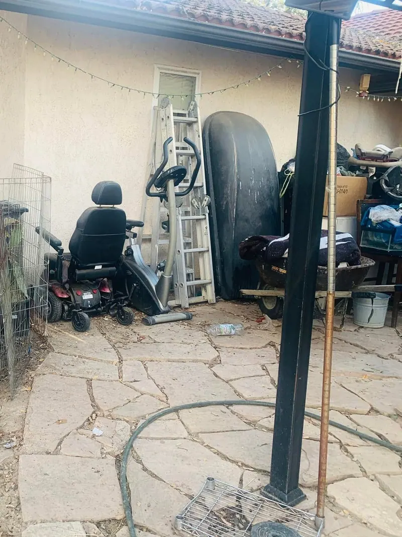 Wire crates; a broken chair; shelving and cut branches stacked along the side of a house